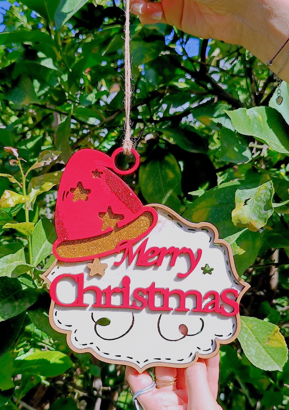 Hand holding Merry Christmas Santa ornament outdoors with green leaves background