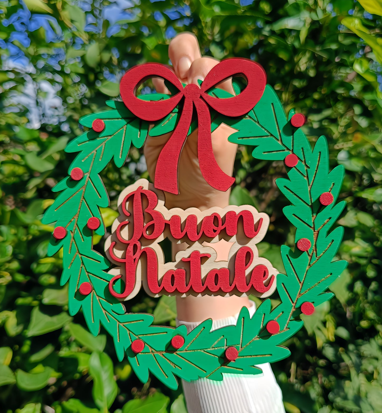 Hand holds a wooden Buon Natale Christmas wreath with red bow, outdoors with green leaves