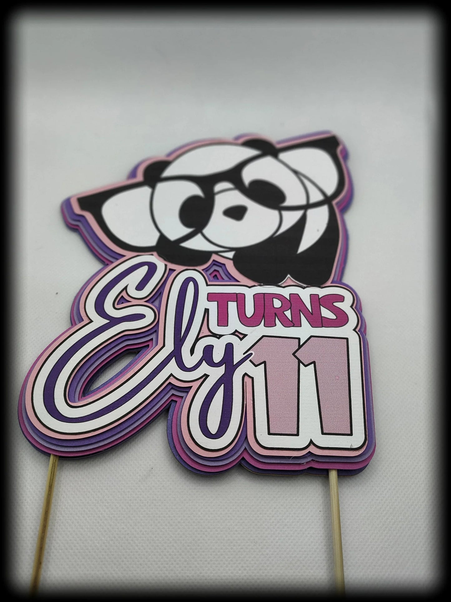 Panda-themed birthday cake topper with 'Ely turns 11' in pink and purple accents