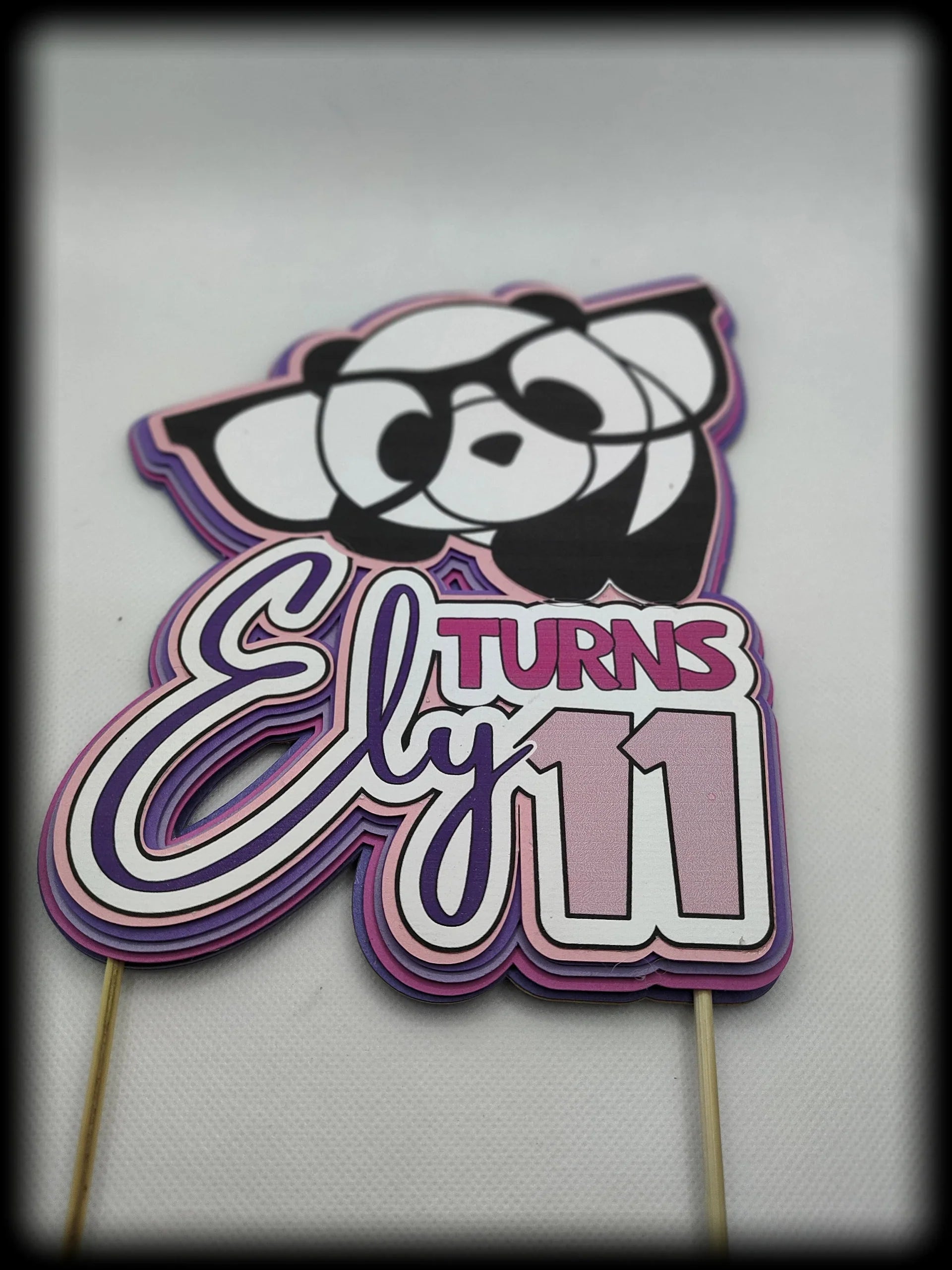 Panda-themed birthday cake topper with 'Ely turns 11' in pink and purple accents