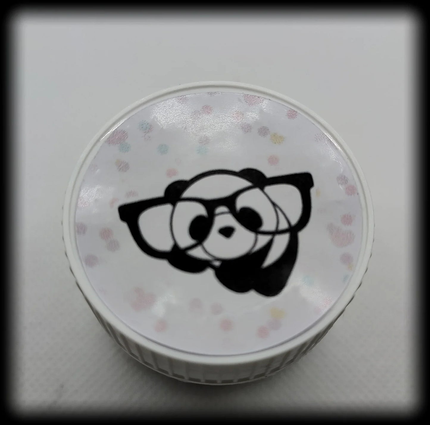 White round container lid with cartoon panda face in glasses and pastel polka dots on top.