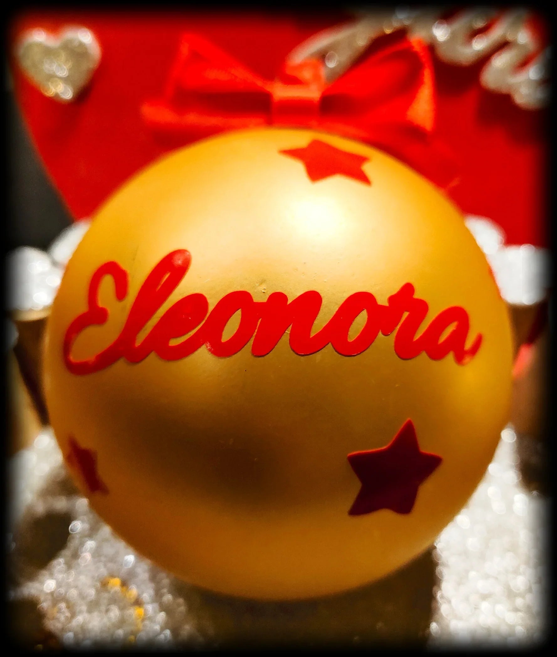 Gold Christmas ornament with red stars and 'Eleonora' text, red bow in background