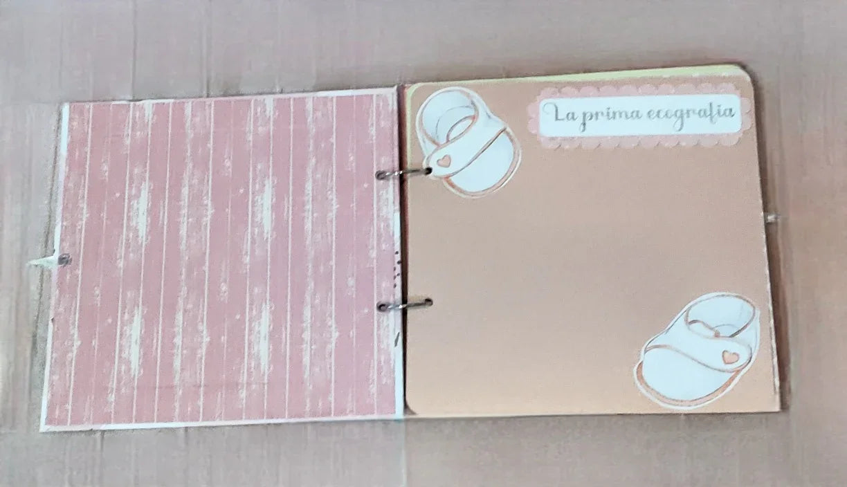 Baby scrapbook page with pink wood pattern, beige page, white baby booties, La prima ecografia text.