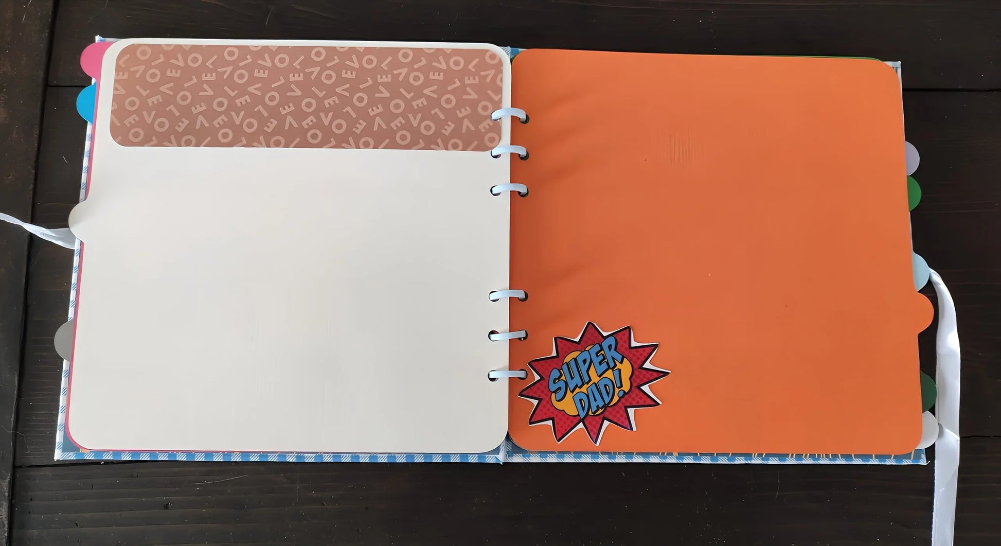Open scrapbook with a blank page and orange 'Super Dad!' comic page on wooden table