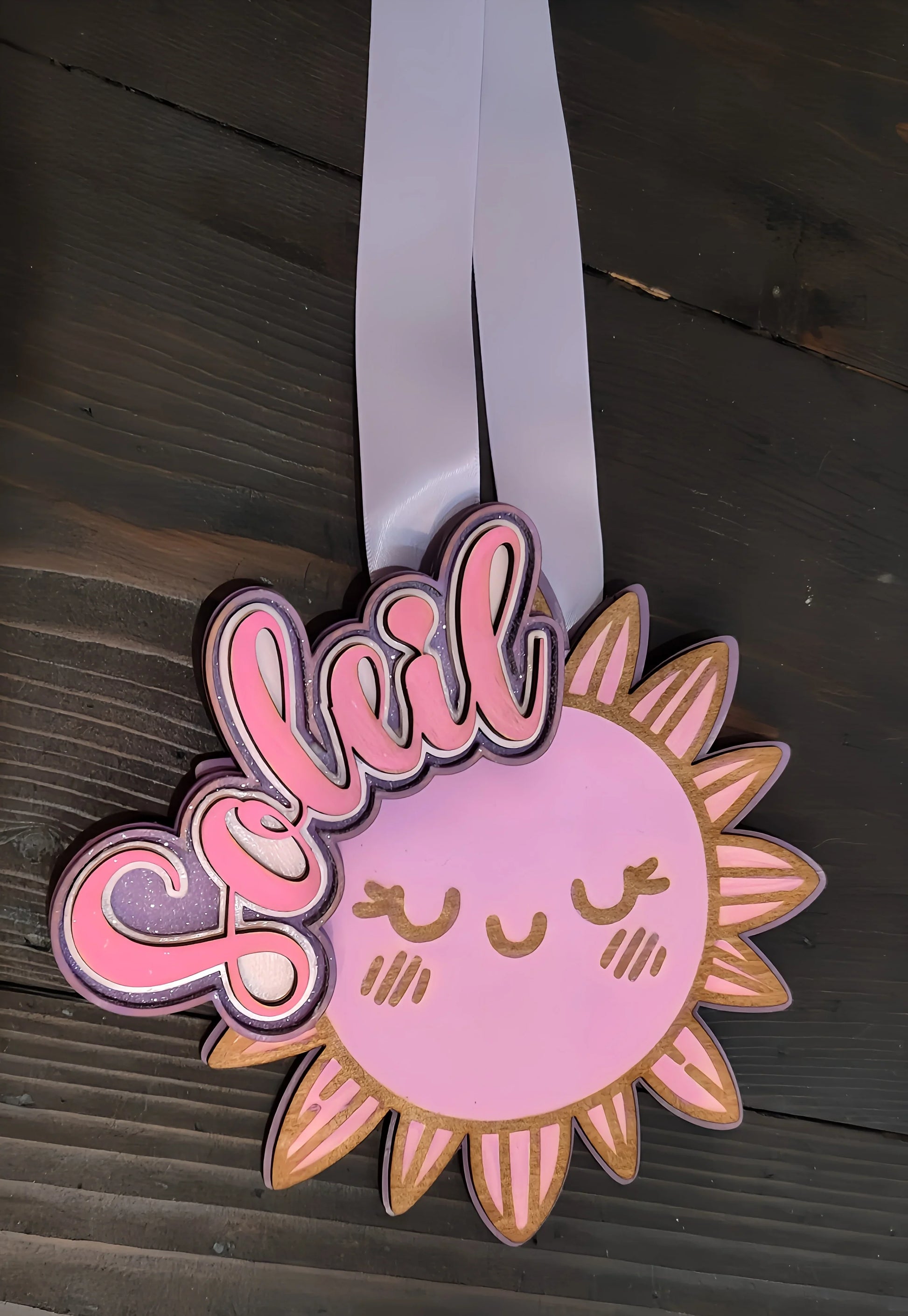 Cute pink sun with closed eyes, cheeks, and 'Soleil' text, wooden background, ribbon badge