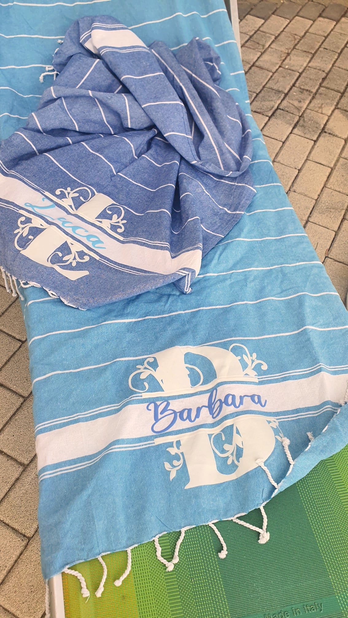 Blue striped personalized beach towels with 'Barbara' and 'Luca' text on poolside chair