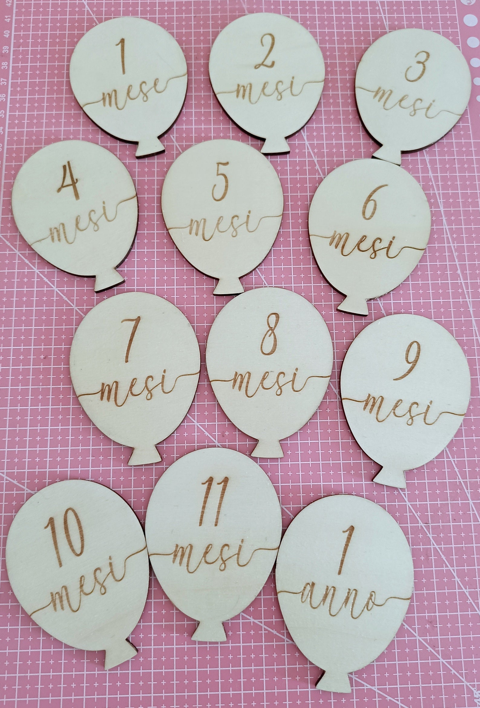 Wooden balloon-shaped monthly milestone markers in Italian on a pink grid background