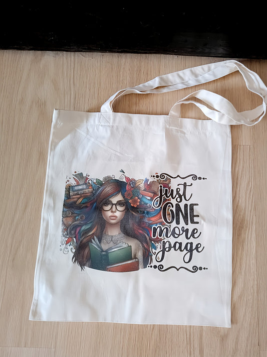 White tote bag with illustrated girl, books, and 'just one more page' text on wood floor