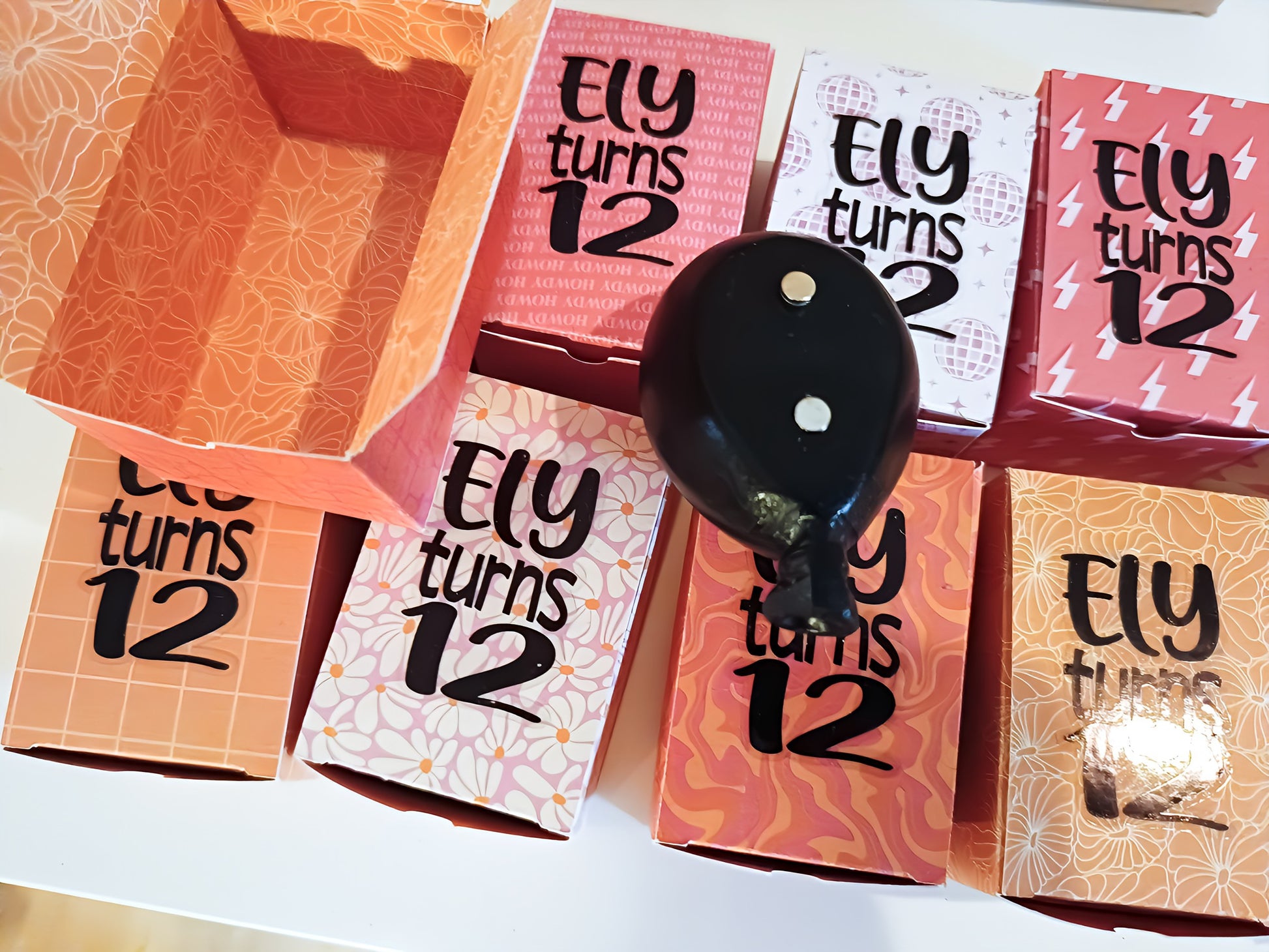 Colorful birthday favor boxes with 'Ely turns 12' text and a black balloon