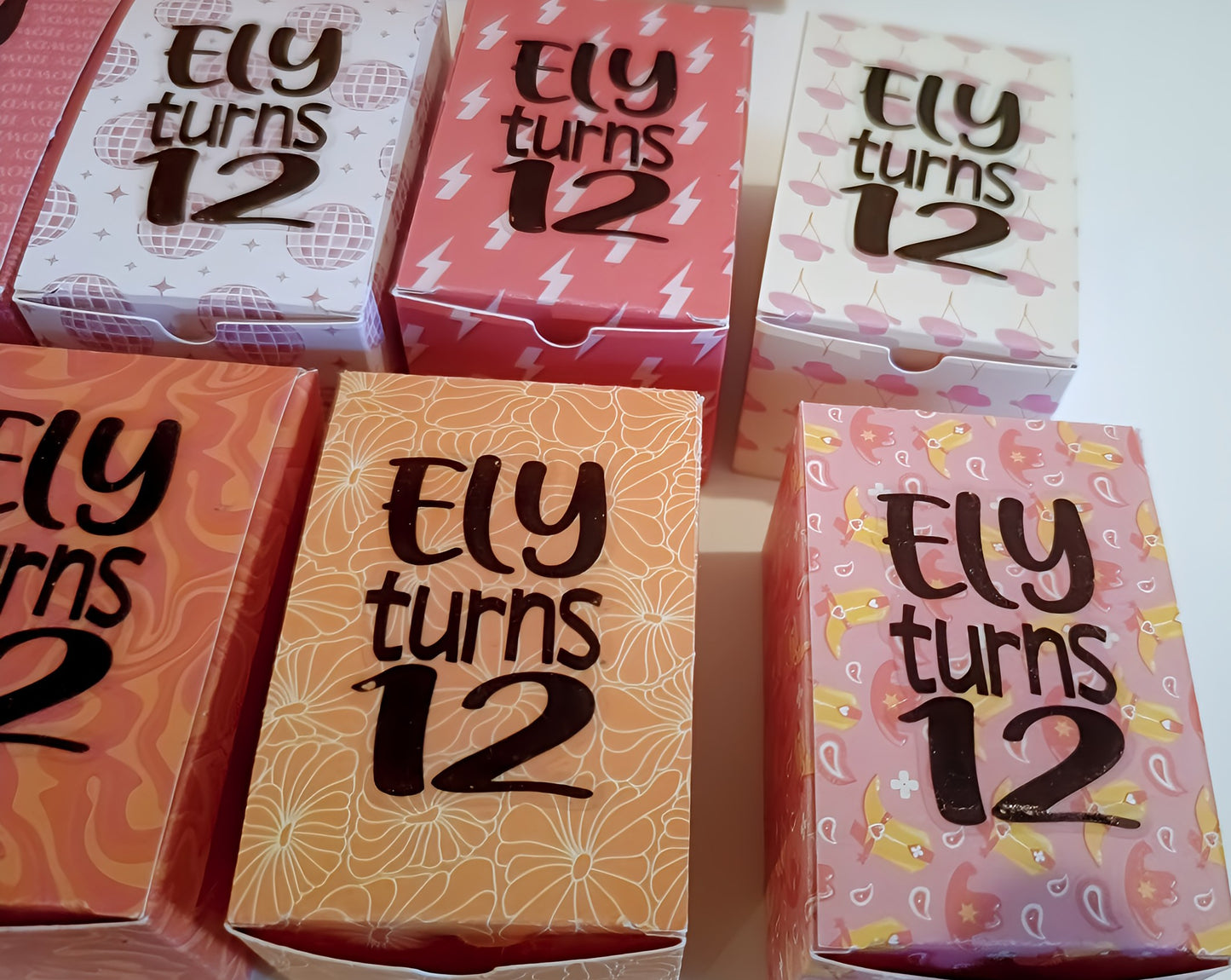Colorful birthday party favor boxes with 'Ely turns 12' in fun themed patterns.
