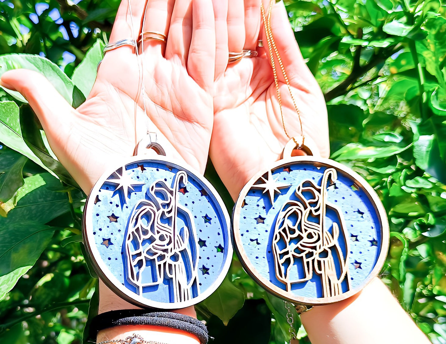Hands holding two round Nativity Christmas ornaments with a blue starry background outdoors