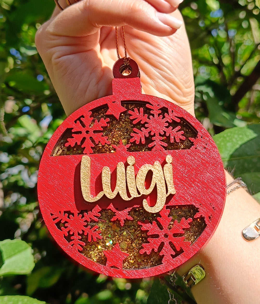 Hand holding red personalized Christmas ornament with 'Luigi' and glitter snowflakes outdoors