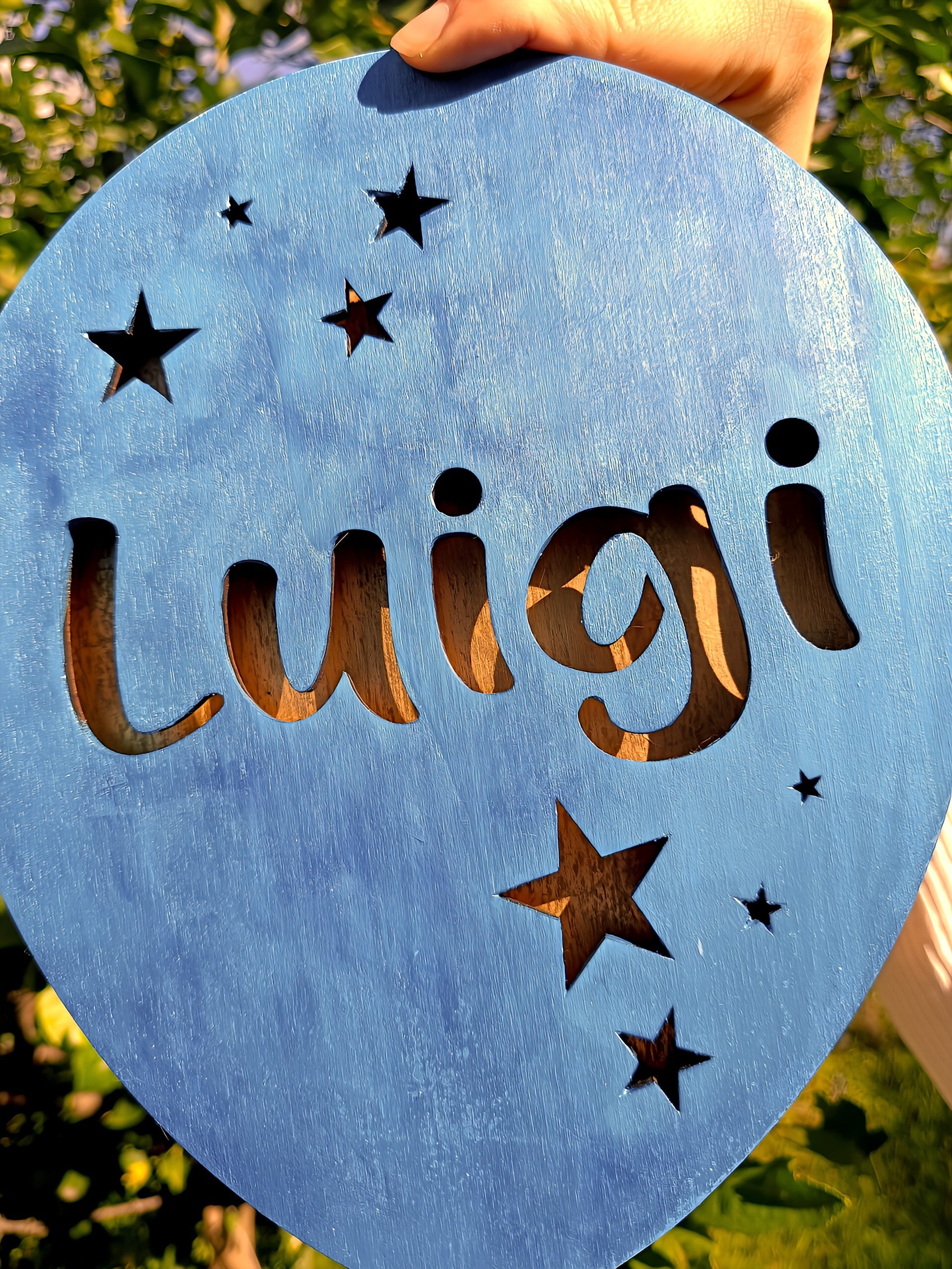 Blue wooden sign with 'Luigi' and star cutouts, held outdoors in sunlight