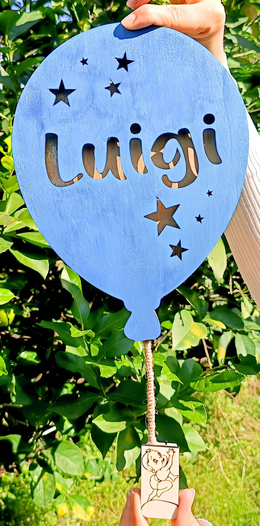 Blue wooden balloon sign with cut-out stars and 'luigi' name, held outdoors in greenery