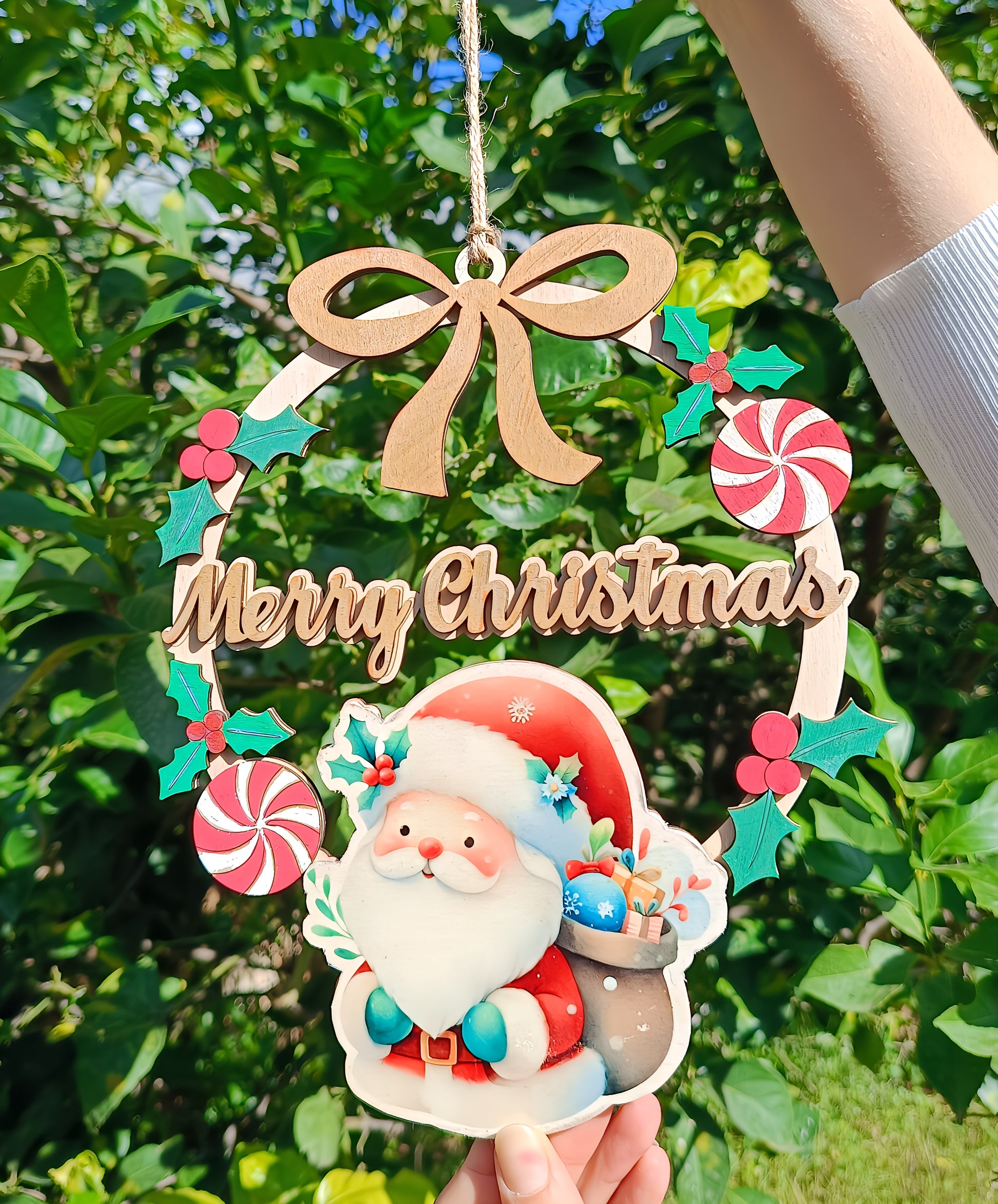 Hand holding a wooden Merry Christmas sign with Santa Claus, ribbon, and candy outdoors