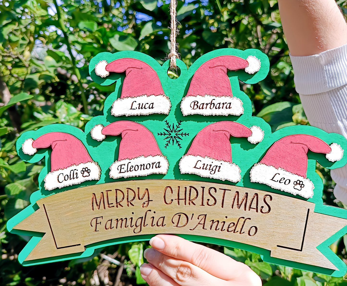 Hand holding personalized wooden Christmas sign with Santa hats and family names, outdoor background