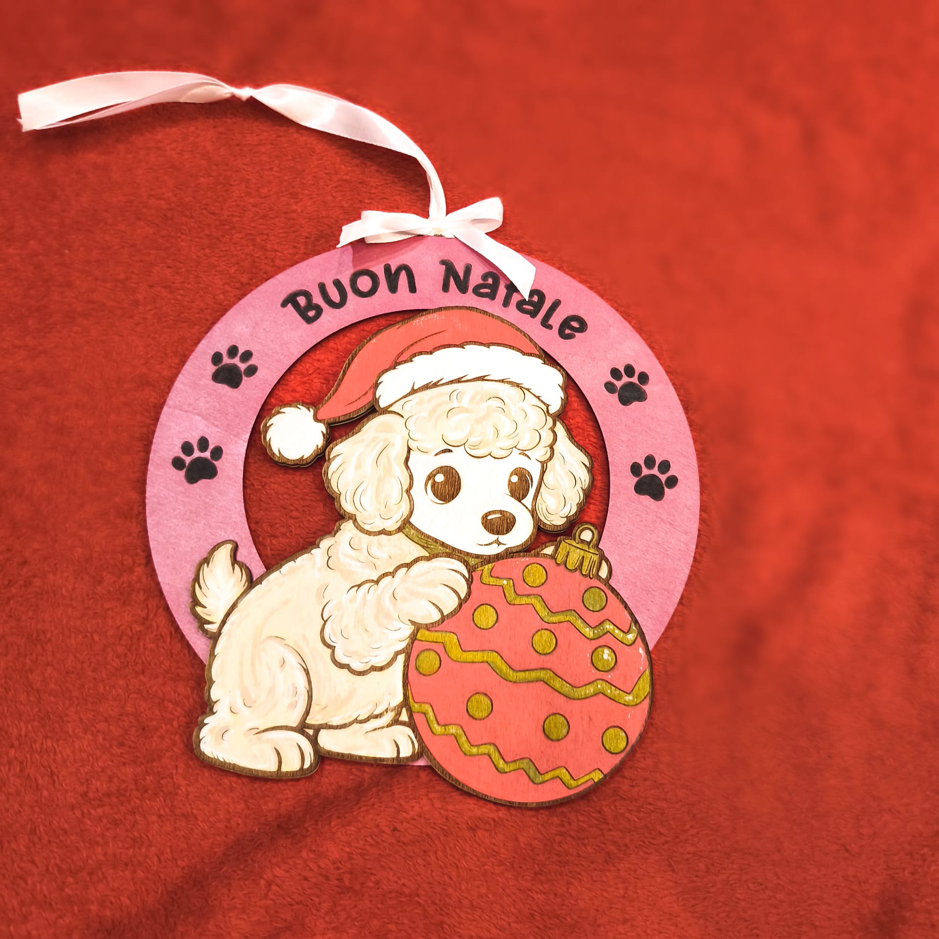 Addobbo natalizio artigianale in legno con barboncino bianco, cappello rosso di Babbo Natale e palla di Natale decorata. Scritta Buon Natale su cornice rosa con impronte di zampe. Nastro bianco per appendere. Decorazione handmade italiana.