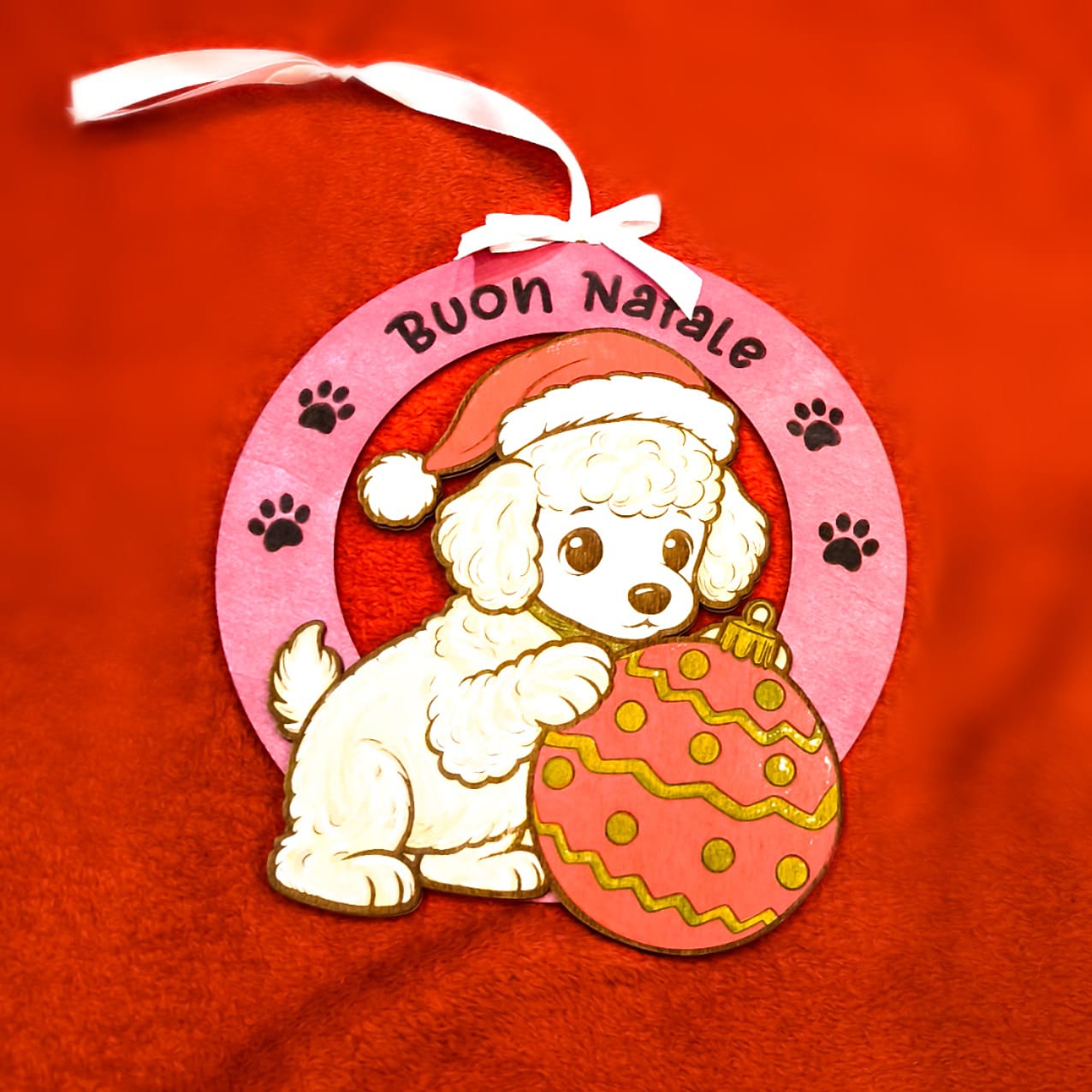 Addobbo natalizio artigianale in legno con barboncino bianco, cappello rosso di Babbo Natale e palla di Natale decorata. Scritta Buon Natale su cornice rosa con impronte di zampe. Nastro bianco per appendere. Decorazione handmade italiana.