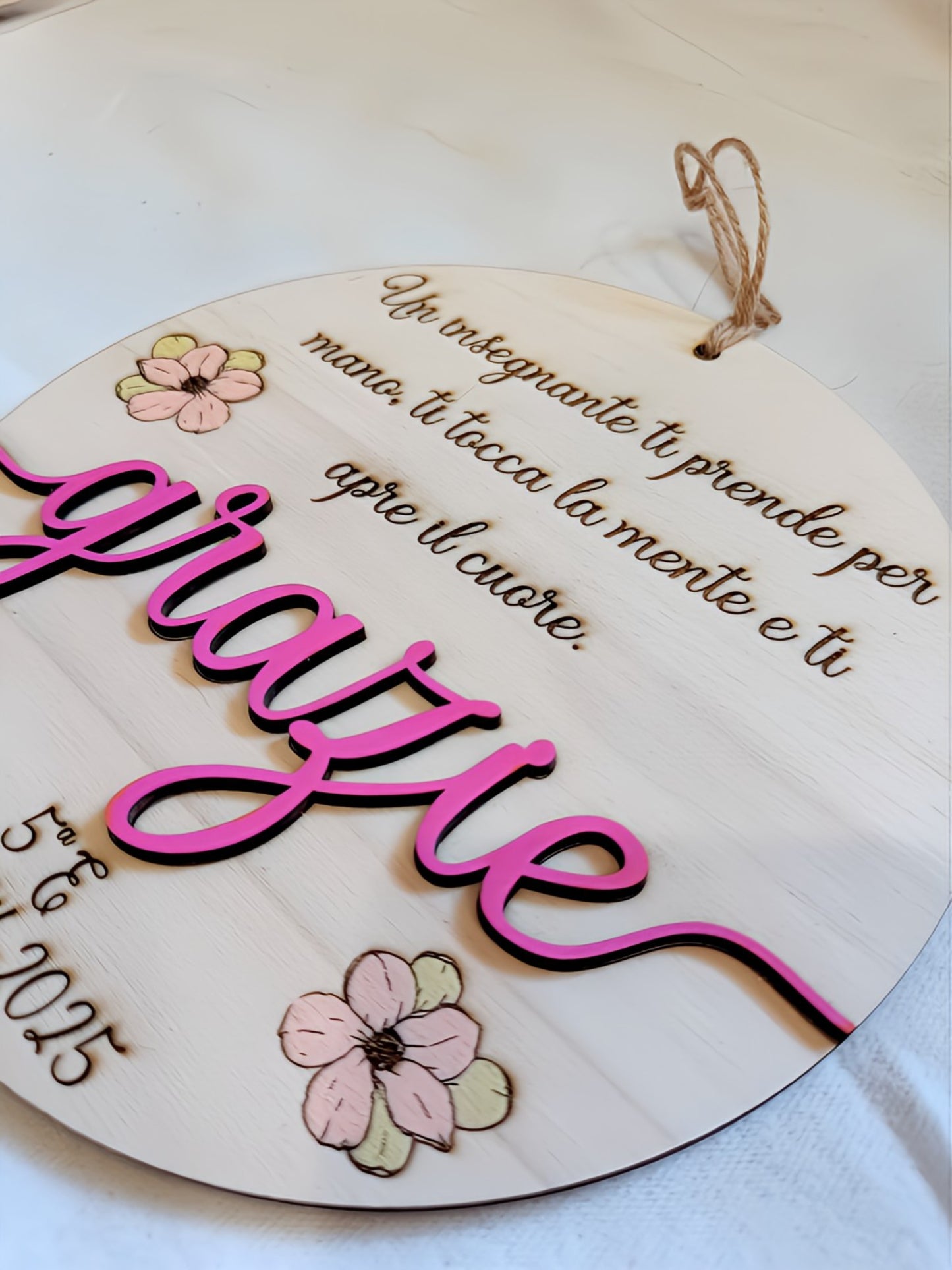 Dettaglio ravvicinato della targa in legno per maestra con scritta tridimensionale "grazie" in legno tagliato e dipinto fucsia, incisione a pirografia della frase motivazionale, fiorellini dipinti a mano in toni pastello e cordino in iuta.