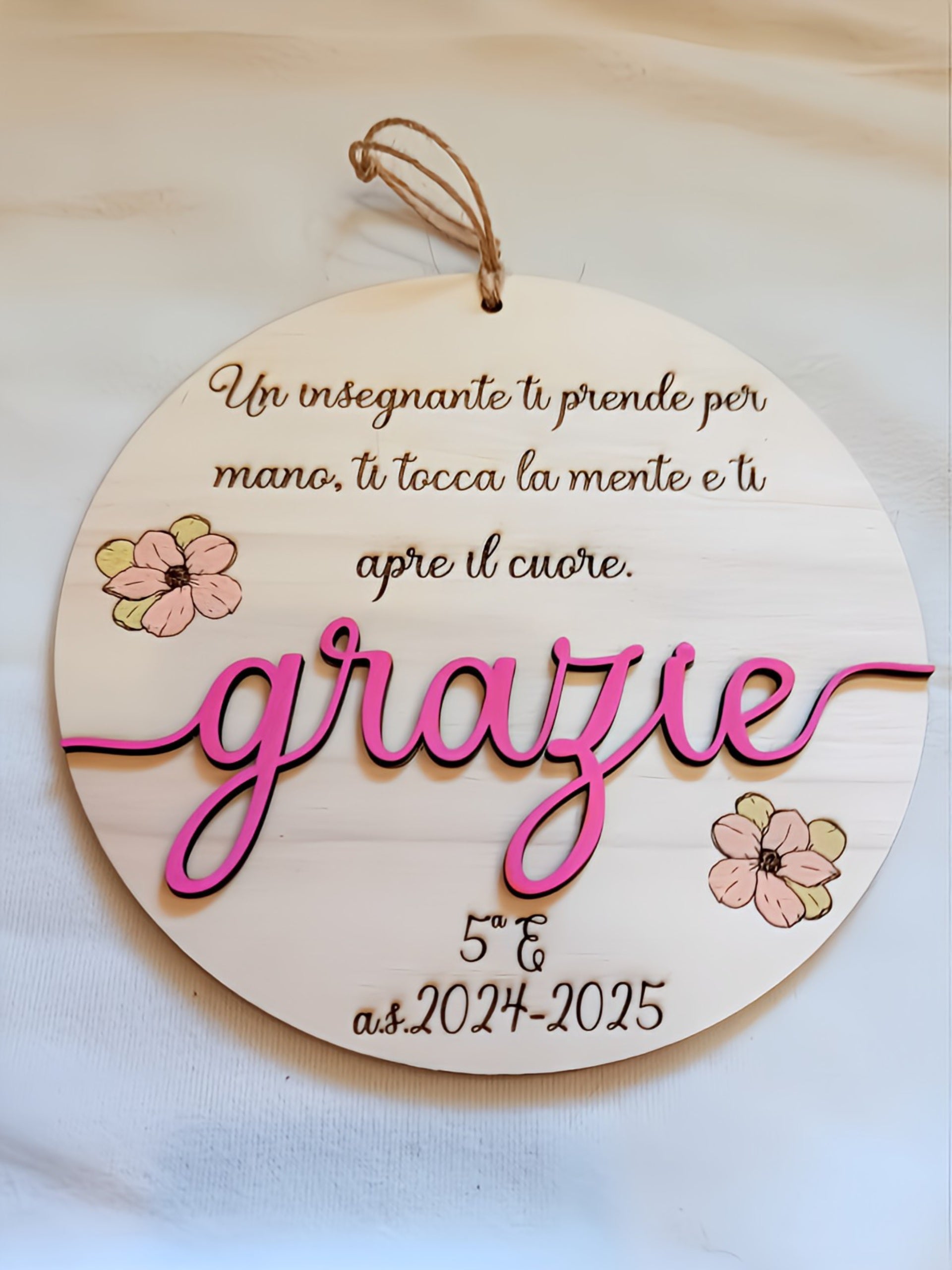 Targa rotonda in legno naturale con incisione a fuoco della frase "Un insegnante ti prende per mano, ti tocca la mente e ti apre il cuore", scritta tridimensionale "grazie" in legno dipinto fucsia, fiori decorativi dipinti a mano in rosa e verde, personalizzazione classe 5ª E anno scolastico 2024-2025 e cordino in iuta.
