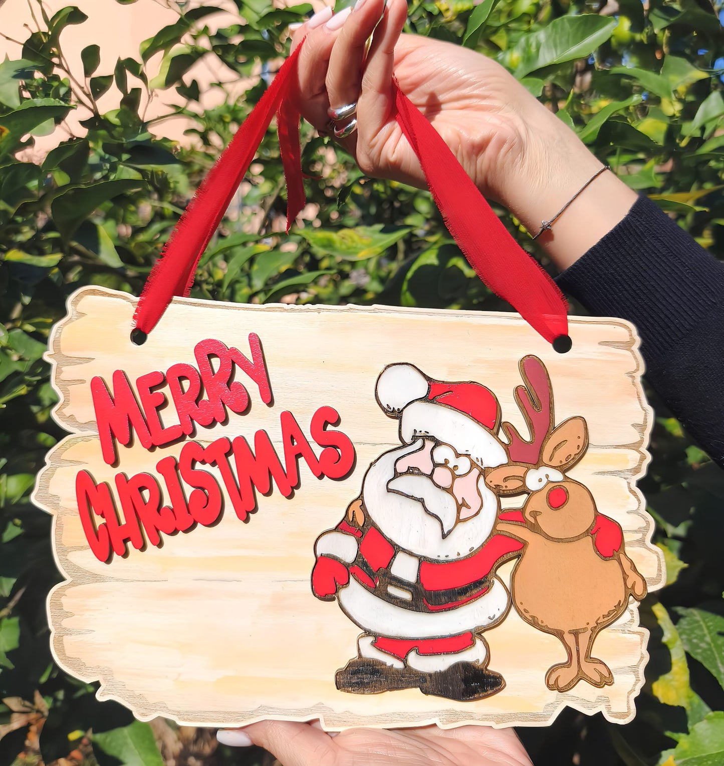 Hand holding wooden Merry Christmas sign with cartoon Santa and reindeer in garden