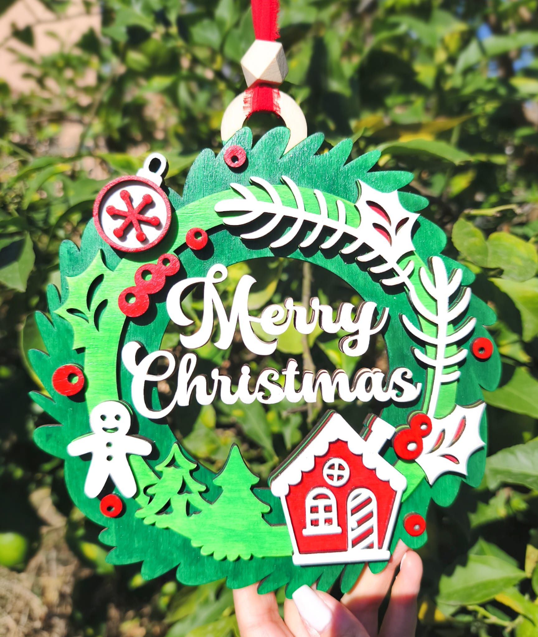 Wooden Merry Christmas wreath ornament with house and gingerbread, held outdoors in sunlight.