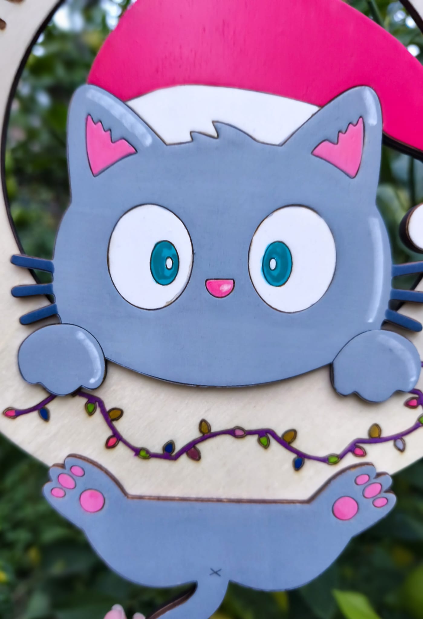 Cute gray cartoon cat with a Santa hat and blue eyes, holding a wreath with Christmas lights